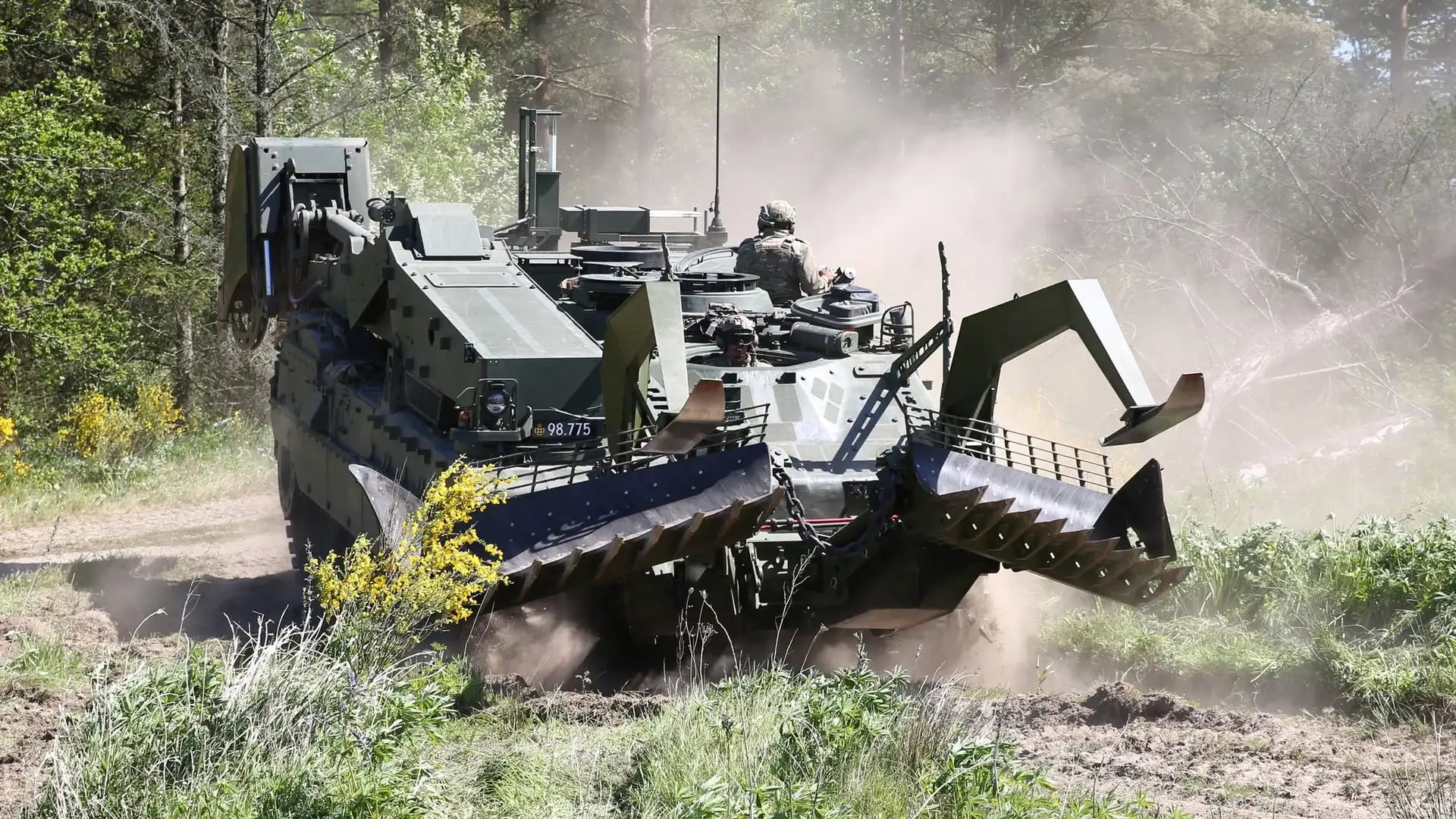 Militært ingeniørkøretøj kører på en jordvej i skovområde og hvirvler støv op.