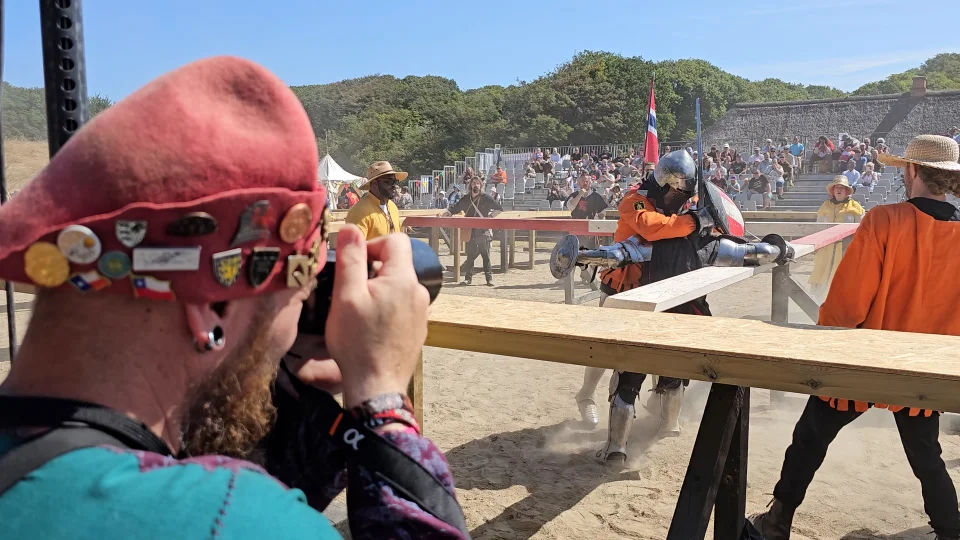 To riddere kæmper i arenaen, mens en væbner springer bagud for ikke at blive ramt. En fotograf i middelalderkostume tager et billede af øjeblikket. Foto: Ditte Haaning, Skive Kommune