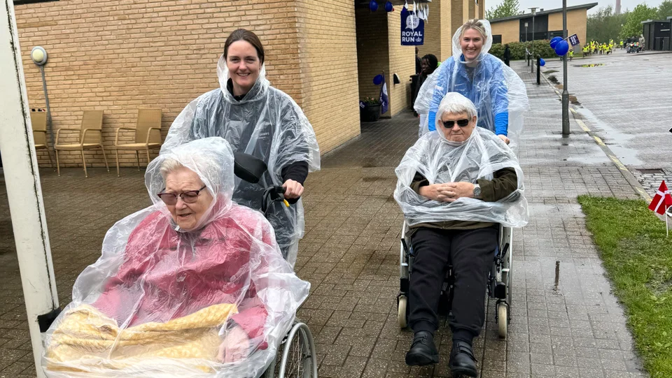 To ældre borgere og to unge mennesker som skubber dem i kørestole til Royal Side Run på Plejecenter Gammelgaard. Alle har regnslag på.