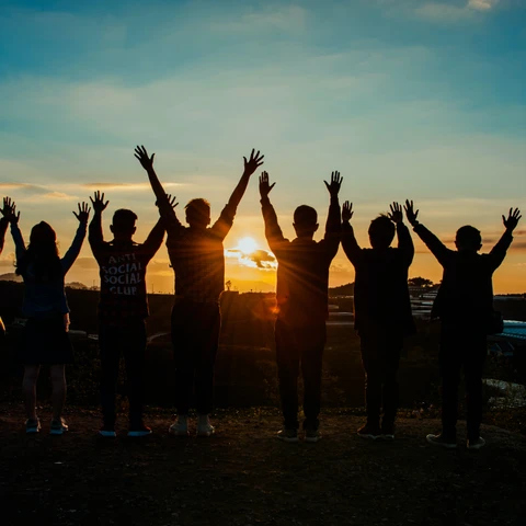 En gruppe mennesker i silhouet, der står og ser mod en solnedgang, med armene strakt over deres hoveder.