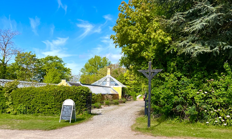 Et skilt står foran en hæk ved en grusbelagt indkørsel. Bag hækken kan man se et drivhus, omgivet af træer og planter. 
