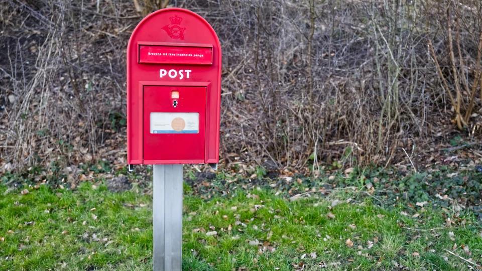 Billede af en rød postkasse, der står frit på et stykke græs i kanten af en vej