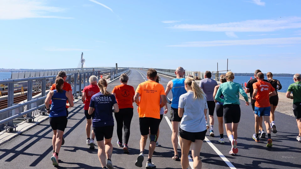 En gruppe mennesker iført sportstøj løber over en bro i solskinsvejr.